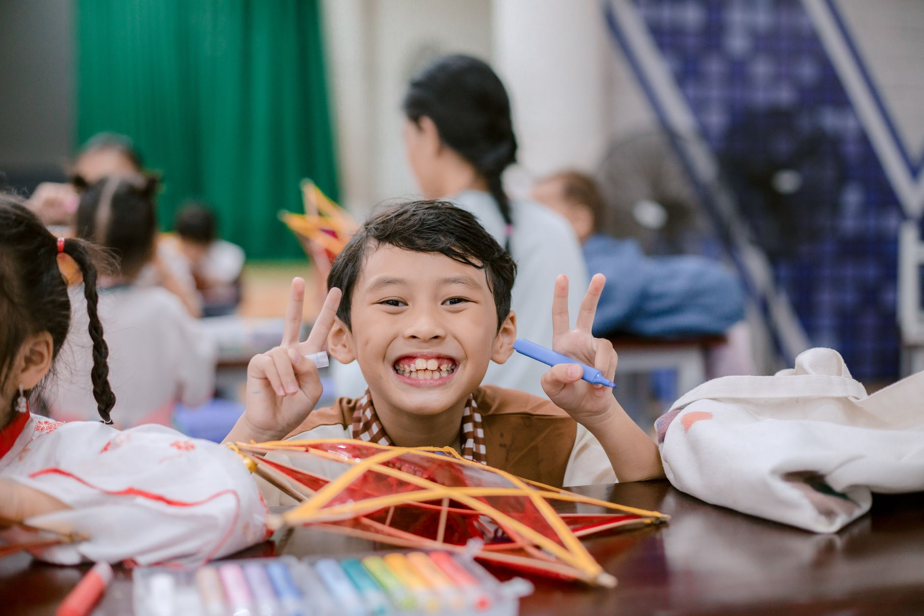Smiling child holding peace sign with colorful pens and art project in classroom setting.