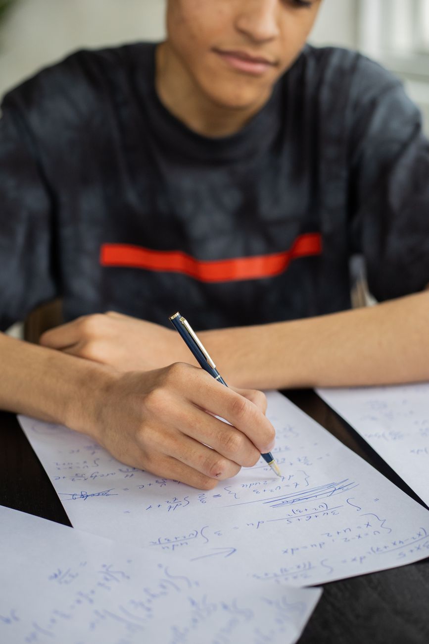 Unrecognizable African American male taking notes on papers and solving equations while sitting at table during studying math lesson in classroom