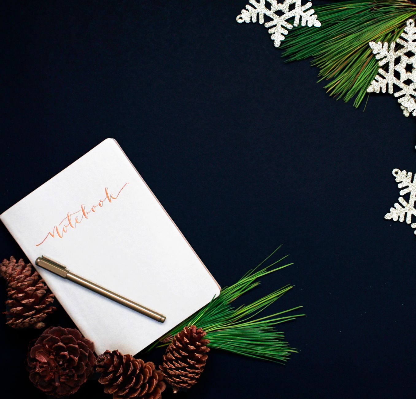A holiday-themed flat lay featuring a notebook, pinecones, and decorative snowflakes against a dark background.