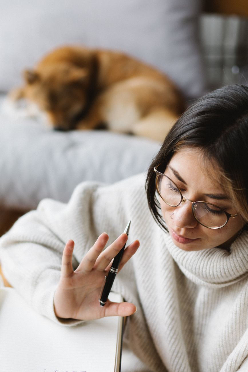 High angle of crop focused young female student with warm sweater and eyeglasses thinking while doing homework assignment at home near sleeping adorable Akita Inu dog