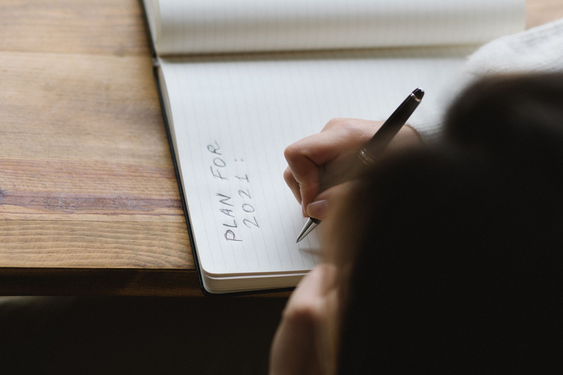 From above of crop unrecognizable female with dark hair sitting at wooden table and writing plan for 2021 in notebook