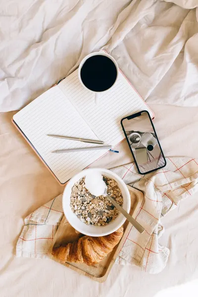 Top view of cozy breakfast setup with coffee, notebook, and smartphone on a bed.