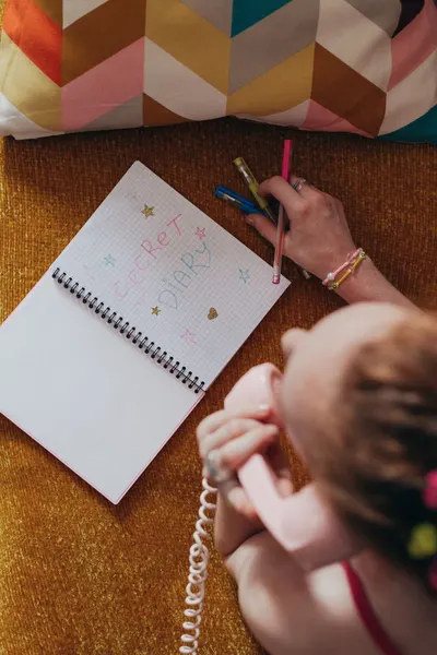 Overhead view of a woman writing in a diary with colored pens while talking on a pink telephone.