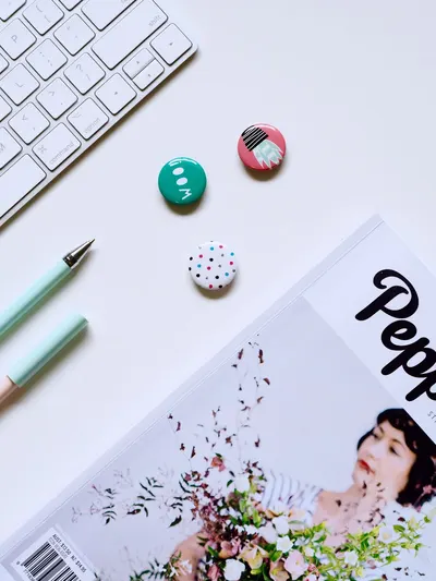 Flat lay of a contemporary desk setup with keyboard, pens, buttons, and a magazine on a white background.