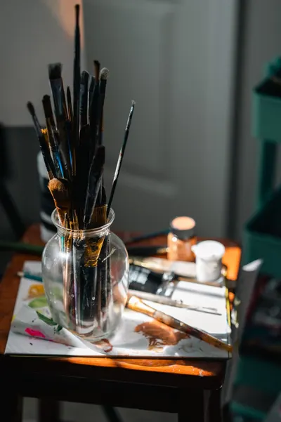 A sunlit art workspace featuring brushes in a glass jar and paint supplies on a wooden table.
