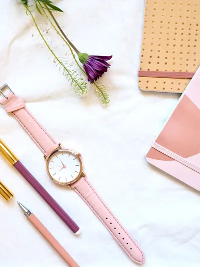 Flat lay of a pink watch, pens, a flower, and notebooks on a white background, showcasing elegance and style.