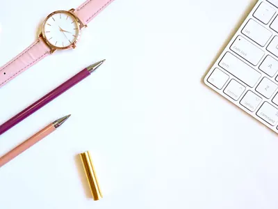 A clean, stylish workspace with a pink watch, colorful pens, and a keyboard on a white background.