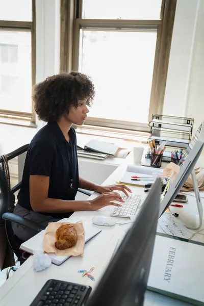 Focused woman typing on keyboard at office desk with computer monitor and documents.