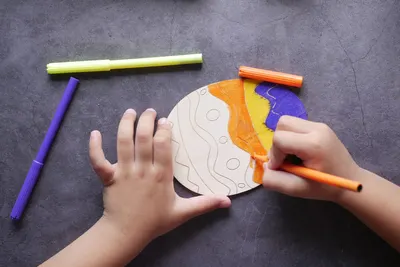 A child's hands coloring a wooden planet cutout using colorful markers on a table.