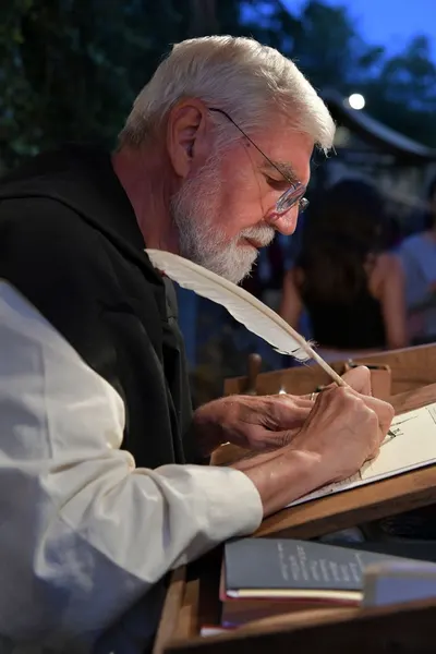 Elderly man using quill and ink to write outdoors at night in Monzambano, Italy.