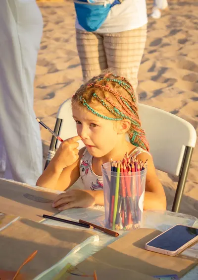 Child with colorful braided hair drawing at an outdoor beach setting.