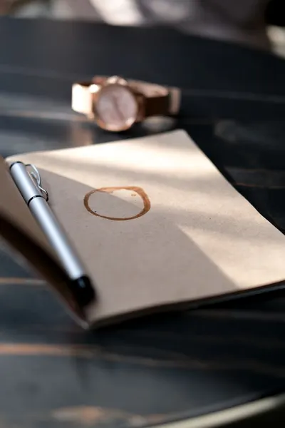 Close-up of a notebook with a coffee stain and pen on a dark table, in soft sunlight.