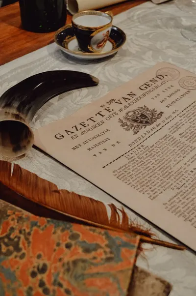 Still life featuring a vintage newspaper, quill, and coffee cup on a wooden table, evoking a historical ambiance.