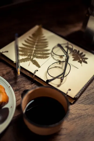 Top view of a notebook with pressed leaves, coffee and eyeglasses on a wooden desk.