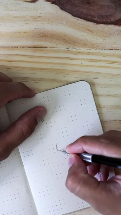 Hands writing with a pen in a dotted notebook on a wooden table, captured from above.