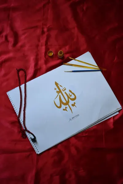 Exquisite Islamic calligraphy on paper with paintbrushes and beads on red fabric background.