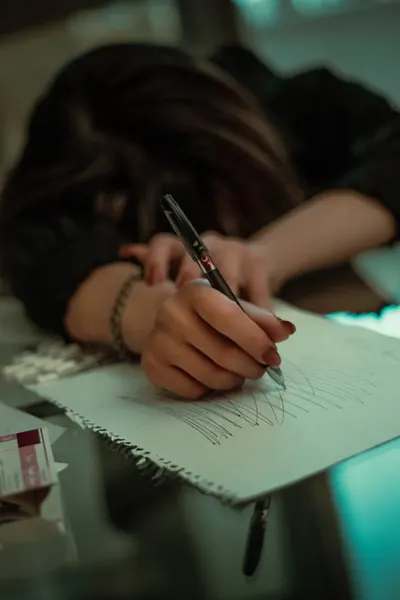 A weary woman leans over a desk scribbling on paper, embodying fatigue and exhaustion.