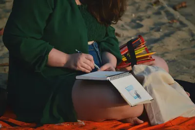 A woman relaxes on a beach while sketching in a notepad, enjoying a creative moment.