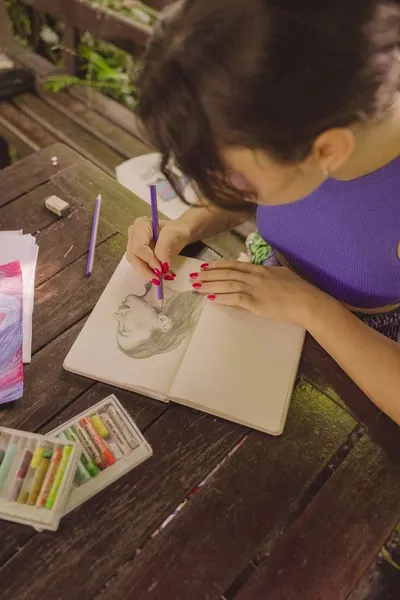 A woman sketching a portrait in a notebook with colored pencils on a wooden table outdoors.
