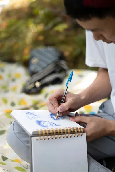 A focused artist sketching portraits with a pen in an outdoor setting on a sunny day.