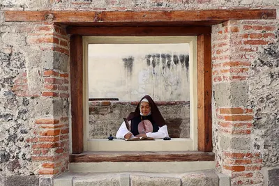 Nun using a quill pen, writing at a window in a historical stone building.