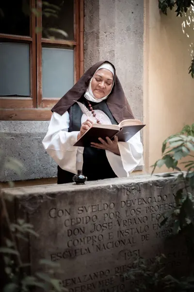 A nun in traditional habit writing in a notebook beside a stone plaque outdoors.