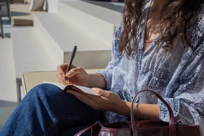 A woman in a blue blouse is writing in a notebook while sitting on outdoor steps, capturing a peaceful moment.