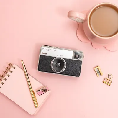 Flat lay of a vintage Kodak camera with pink stationery and coffee on a pink desk.