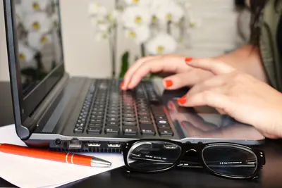 Close-up of a woman's hands typing on a laptop with glasses and a notepad on the desk.