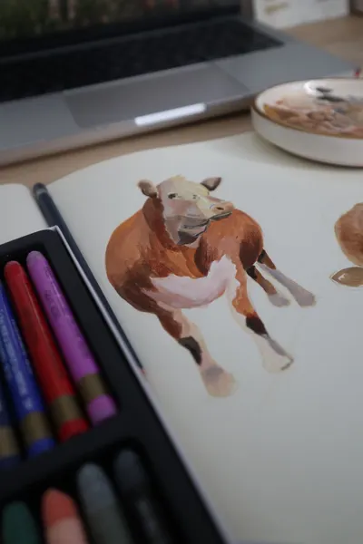 A detailed watercolor cow sketch in a notebook with colored pencils nearby.
