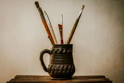 A collection of paintbrushes in a rustic ceramic jug placed on a wooden table, perfect for creative projects.