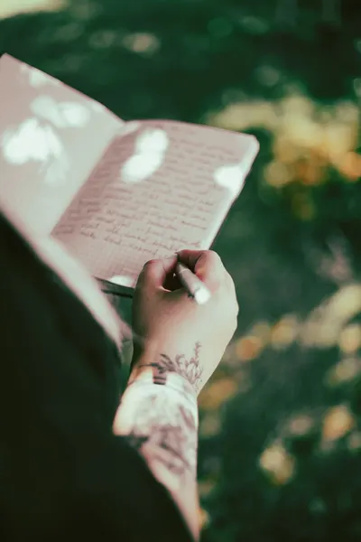 Close-up of a tattooed arm writing in a journal amidst natural lighting outdoors, capturing creativity and reflection.