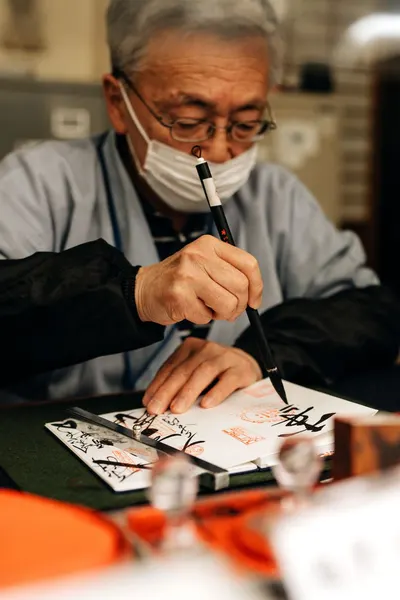 An elderly artist practicing traditional Japanese calligraphy indoors, demonstrating skilled brushwork and cultural craftsmanship.