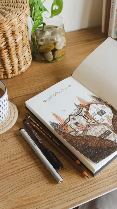 A cozy creative setup featuring a sketchbook, pencils, a plant, and a coffee mug on a wooden desk.
