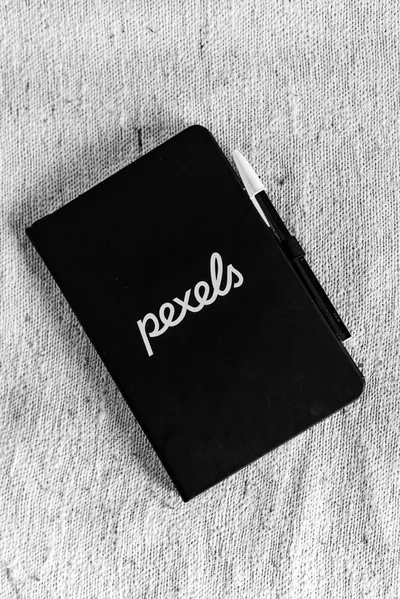 Monochrome image of a closed black notebook with a pen placed on textured fabric background.