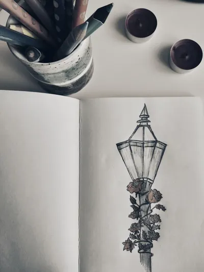 A sketchbook page with a detailed street lamp drawing surrounded by roses, accompanied by colored pencils and candles.