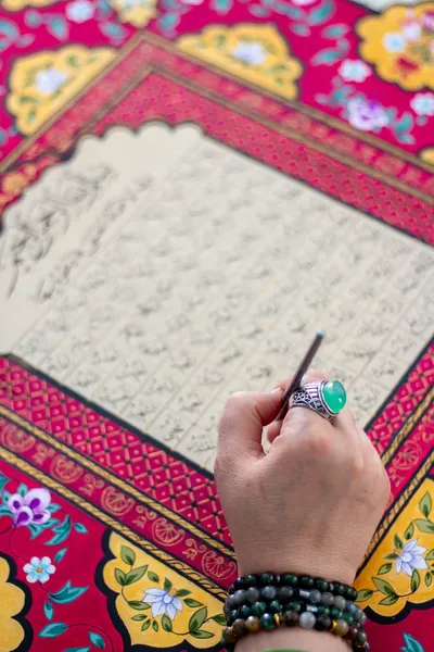 A hand with a ring elegantly performs calligraphy on ornate, colorful fabric.