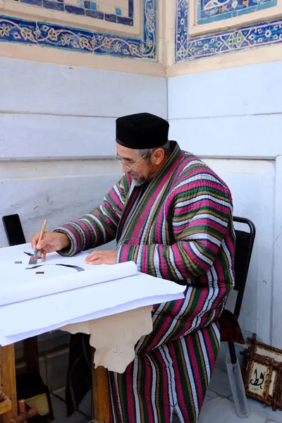 A man in traditional Uzbek attire practicing calligraphy at an outdoor setting.