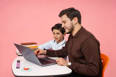 Father guiding his son with online learning on a laptop against a pink background.