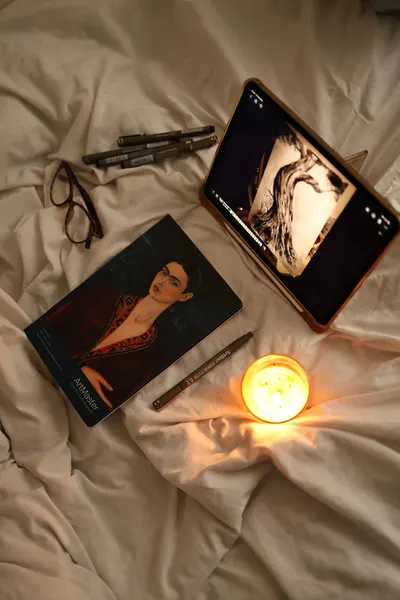 An artistic setup featuring a tablet displaying art, book, and candle on a cozy bed setting.
