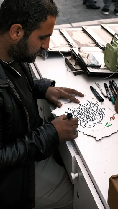 A man carefully designs calligraphy on paper at a street-side workspace, surrounded by art tools.