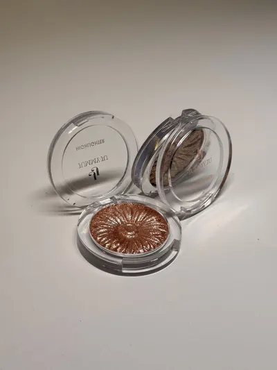 Open and closed containers of shimmering makeup highlighter on a neutral background.