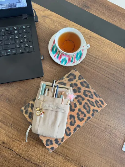 Flat lay of a leopard print notebook, pens, tea, and laptop on a wooden desk.