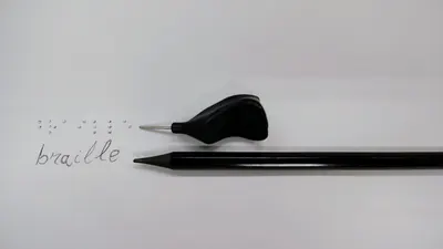 Close-up of a stylus and slate with Braille writing on white surface.