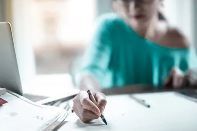 Woman concentrating on sketching at a desk with a laptop and papers in an indoor office setting.