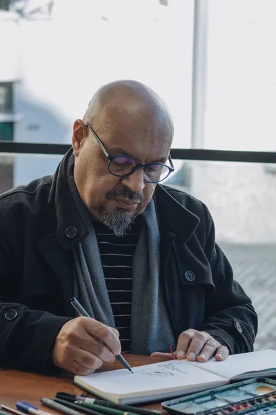 Middle-aged man sketching in a notebook by a window, wearing glasses and a coat.