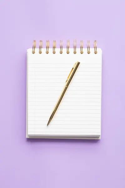 A gold pen resting on a blank lined spiral notebook with a purple background, ideal for stationery enthusiasts.