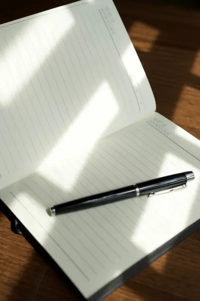 A black pen rests on an open notebook in a sunlit workspace, casting shadows.