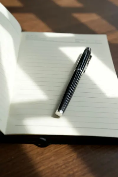 A black pen elegantly rests on a lined open notebook in natural light.