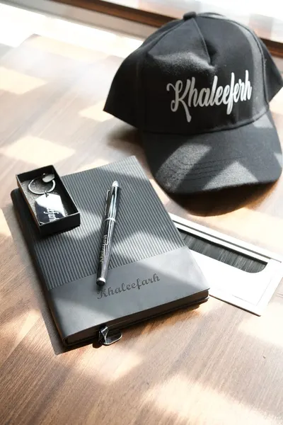Elegant black cap and notebook with pen on a sunlit wooden table, offering a minimalist and stylish look.